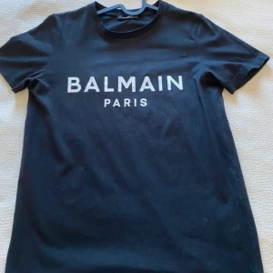 Svart t-shirt från Balmain Paris - Snygg svart t-shirt från Balmain Paris med vit logga tryckt på bröstet. Klassisk rund halsringning och korta ärmar. Perfekt för en stilren och trendig look.