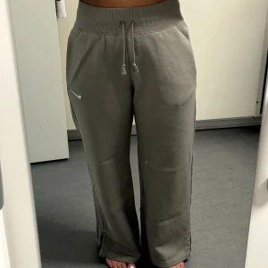 Grå mjukisbyxor från Nike - Gråa mjukisbyxor från Nike med bred resår i midjan och dragsko. Byxorna har raka, vida ben och en liten vit Nike-logga på ena låret. Perfekta för en avslappnad stil. Oversize