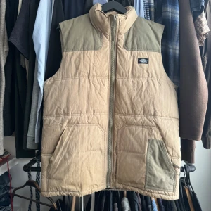 Beige quiltad väst från Dickies - Säljer en beige quiltad väst från Dickies med gröna axelpartier och dragkedja framtill. Västen har två stora fickor och en hög krage. Perfekt för lager-på-lager och har en klassisk streetwear-look.