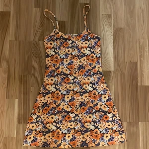 Blommig sommarklänning  - Säljer en blommig klänning från H&M i storlek XS. Klänningen har smala axelband och är ärmlös med ett färgglatt blommönster i orange, lila och vitt. Perfekt för varma dagar och har en lätt utsvängd passform. Aldrig använd!🧡