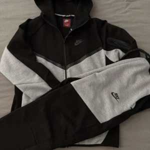 Svart och grå zip-hoodie från Nike - Säljer en stilren svart och grå zip-hoodie från Nike med huva och dragkedja. Jackan har lång ärm, diskret Nike-logga på bröstet och en ficka med dragkedja på ärmen. Perfekt för en sportig look.