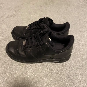 Svarta Nike Air Force 1 sneakers - Säljer ett par helsvarta Nike Air Force 1 sneakers med klassisk design. Skorna har snörning, rund tå och platt sula. Tillverkade i skinn med diskret Nike-logga på sidan och metallbricka på snörena. Perfekta för en stilren och clean look.