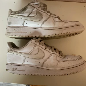 Vita Nike Air Force 1 sneakers - Klassiska vita Nike Air Force 1 sneakers med lågt skaft och snörning. Skorna har en rund tå och platt sula, samt den ikoniska swoosh-loggan på sidan. Perfekta för en streetwear-look och passar till det mesta.