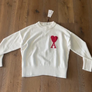 Vit stickad tröja från Ami Alexandre Mattiussi - Vit stickad tröja med rund hals och ribbade muddar från Ami Alexandre Mattiussi. På bröstet finns ett rött hjärta och bokstaven A. Tröjan har en avslappnad passform och är perfekt för en stilren look.