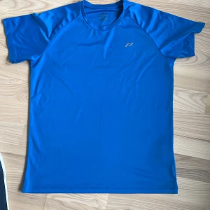 Blå träningst-shirt från Pro Touch - Säljer en blå kortärmad träningst-shirt från Pro Touch. T-shirten är tillverkad i ett syntetmaterial som andas och har en enkel, sportig design med liten logga på bröstet. Perfekt för träning eller sportaktiviteter.