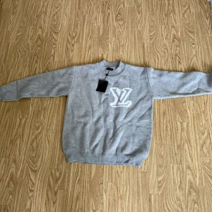 Grå stickad tröja med LV-logga - Säljer en grå stickad tröja från Louis Vuitton med vit LV-logga både fram och bak. Tröjan har rund halsringning och långa ärmar. Perfekt för dig som gillar stilrena och exklusiva plagg.