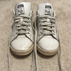 Vita Adidas Stan Smith sneakers - Klassiska vita Adidas Stan Smith sneakers med blå detalj på hälen. Skorna är väl används vilket syns tex på skosnörets färg och andra ställen. Storlek 40, skriv för mer info💕