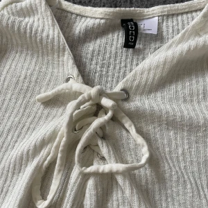 Vit ribbad tröja med snörning från H&M - Säljer en vit ribbad långärmad tröja från H&M med snygg snörningsdetalj framtill vid halsen. Perfekt för dig som gillar enkla men trendiga plagg. Supermjuk och skön att ha på sig!