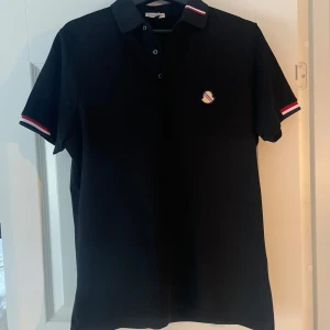 Ny svart moncler piké med rödvita detaljer och tag - Snygg svart pikétröja från Moncler med klassisk krage och knappar och ny med tag. Rödvita ränder på krage och ärmslut samt liten Moncler-logga på bröstet. Perfekt för en stilren look.   Aldrig använd Detaljer: 	•	Storlek: M 	•	Färg: Svart 	•	Material: 100% piqué-bomull 	•	Knappar med “Moncler / Depuis 1952” 	•	Broderad logo på bröstet                   Hör av er vid frågor eller fler bilder. Pris kan diskuteras 