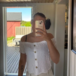 Vit offshoulder blus med knappar - Säljer en vit offshoulder blus med träknappar framtill. Blusen har korta ärmar och en lös passform, perfekt för varma dagar. Materialet är lätt och luftigt, vilket gör den skön att bära.