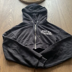 Svart croppad hoodie med tryck - Säljer en svart croppad hoodie med dragkedja och huva. På bröstet och ryggen står det 'OUT OF CONTROL' i vitt tryck. Perfekt för en edgy streetstyle-look.