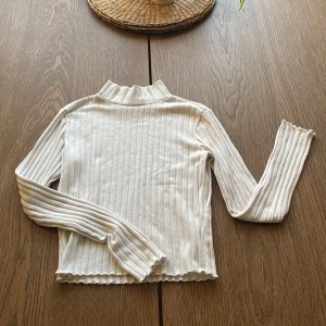 Vit ribbad långärmad topp från Gina Tricot - Säljer en vit ribbad långärmad topp från Gina Tricot i storlek XS. Tröjan har en hög krage och vågiga kanter vid ärmslut och nederkant. Perfekt att styla till jeans eller kjol för en enkel och snygg look.