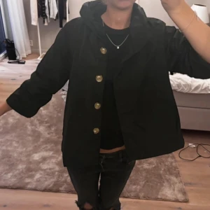 Svart jacka med stora knappar - Jacka i vattentåligt ”prassligt” material, trekvartsarmar och krage. 