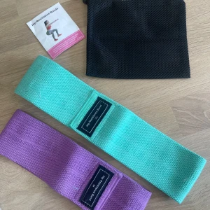 Träningsbälten i mintgrön och lila - Två breda träningsbälten i mintgrönt och lila, perfekta för styrketräning och aktivering av höft och ben. Banden är tillverkade i slitstarkt tygmaterial och kommer med en svart förvaringspåse.