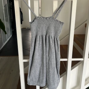 Grå klänning med smock från Juicy Couture - Säljer en grå axelbandsklänning från Juicy Couture i storlek XS. Klänningen har smock upptill och tunna axelband, vilket ger en bekväm och avslappnad passform. Perfekt för varma dagar eller som ett snyggt lagerplagg.