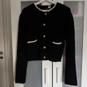 Svart kofta med guldknappar och vita detaljer - Säljer en svart kofta från H&M med guldknappar och vita kanter vid hals, fickor och ärmslut. Koftan har två små fickor framtill och är croppad i modellen. Perfekt för en stilren look!