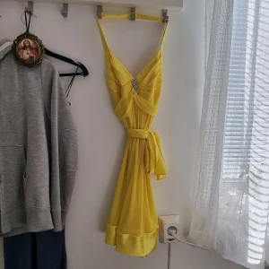 Gul långklänning med strassdetalj - Säljer en gul långklänning med smala axelband, v-ringning och dekorativ strassdetalj framtill. Klänningen har omlottliknande drapering och ett matchande bälte i midjan. Ger en superfin figur