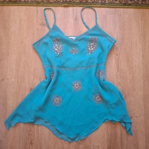 Turkos linnetopp med paljettmönster - Säljer en turkos linnetopp med smala axelband och dekorativa paljettmönster framtill. Toppen har asymmetrisk nederkant och är tillverkad i ett tunt, lätt material som ger en luftig känsla.