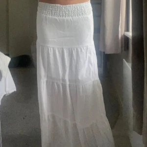 Vit långkjol med smock i midjan - Säljer en vit långkjol med smockad midja och volanger. Kjolen är luftig och har en flowy känsla, perfekt för varma dagar. Passar dig som gillar en enkel men stilren look. Vet ej storleken på den har klippt lappen!!