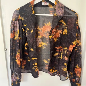 70-tals Svart blommig transparent blus  - Säljer en svart transparent blus med färgstarkt blommönster i orange, gult och lila. Blusen har långa ärmar och knappar.