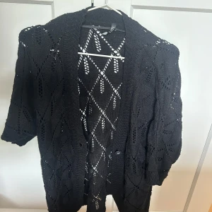 Svart stickad kofta från LMX - Säljer en svart stickad kofta från LMX med korta ärmar och ett genomgående hålmönster. Koftan har en öppen design och är perfekt att slänga över en topp för en avslappnad look.