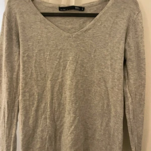 Ljusgrå v-ringad stickad tröja från 157 - En enkel och stilren ljusgrå stickad tröja från 157 med v-ringning. Tröjan har långa ärmar och ribbade muddar vid ärmslut och nederkant. Perfekt för lager-på-lager under kyligare dagar.