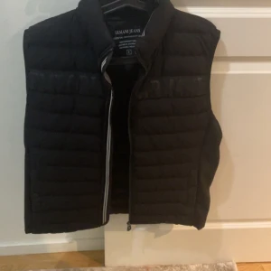 Svart dunväst från Armani Jeans - Säljer en svart dunväst från Armani Jeans med quiltad design och dragkedja framtill. Västen är lätt och har hög krage med logotyp på baksidan. Perfekt för lager-på-lager och enkel att matcha med olika outfits. Utan zip