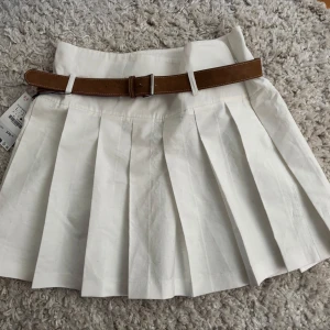 Vit plisserad kjol med brunt bälte - Snygg vit kortkjol med breda plisseringar och ett brunt bälte i midjan. Kjolen har en klassisk och stilren look som passar perfekt till många olika outfits. Bältet ger en extra detalj och markerar midjan på ett snyggt sätt.