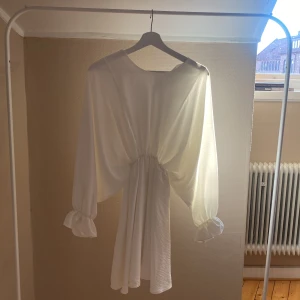 Vit omlottklänning med långa ärmar - Säljer en vit omlottklänning med långa, lösa ärmar och knyt i midjan. Klänningen har en lätt, flowy känsla och v-ringad hals. Perfekt för sommarens tillfällen! Är mer som en storlek S men sitter nog bra på båda.