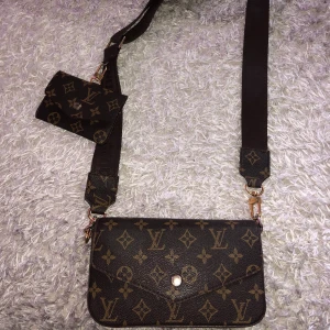 axelväska med monogram - Säljer en brun axelväskamed klassiskt monogrammönster och bred axelrem. Väskan har en mindre avtagbar plånbok och insida i rött tyg med flera kortfack. Perfekt för dig som vill ha både stil och funktion.