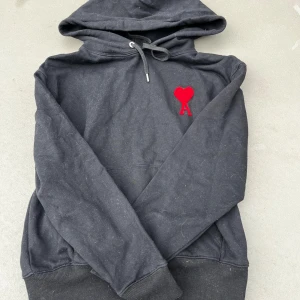 Svart hoodie från Ami Paris med rött hjärta - Svart hoodie från Ami Paris med den ikoniska röda hjärta-A-loggan broderad på bröstet. Tröjan har huva med dragsko och ribbade muddar vid ärmslut och nederkant. Perfekt för en avslappnad och trendig stil.
