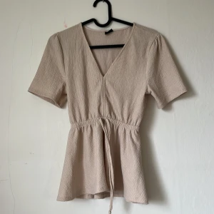 Beige omlottopp från Gina Tricot - Säljer en beige omlottopp från Gina Tricot i storlek XS. Toppen har korta ärmar, v-ringning och knyt i midjan för en snygg passform. Perfekt till både jeans och kjol! Aldrig använd!