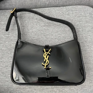 Svart axelväska från Yves Saint Laurent - Säljer en elegant svart axelväska från Yves Saint Laurent i glansigt material med guldigt YSL-emblem framtill. Väskan har justerbart axelband och klassisk design, perfekt för att lyfta vilken outfit som helst. Priset kan diskuteras 