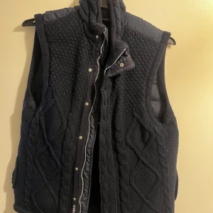 Marinblå  stickad dunväst från J.Lindeberg - Säljer en marinblå dunväst från J.Lindeberg med stickad framsida och vadderad baksida. Västen har dragkedja och knappar framtill samt ett snyggt kabelstickat framsida. Den är i myxket bra skick 