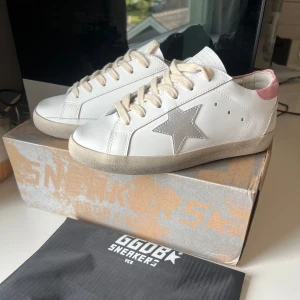 Golden Goose rosa och vita - Säljer ett par vita sneakers från Golden Goose med silverfärgad stjärna på sidan och ljusrosa detalj vid hälen. Skorna har beige snörning och klassisk rund tå. Perfekta för dig som gillar stilrena och trendiga sneakers. Hör av dig innan köp!