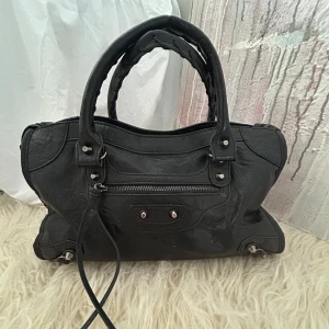 Svart väska - Snygg svart handväska med silverfärgade metalldetaljer och dragkedja framtill. Fint skick! Kontakt mig om du har några frågor. Längd: 40 cm, Bredd: 10 cm, Höjd: 24 cm