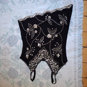 Svart paljettkorsett med blommönster - Supersnygg svart korsett med silvriga paljetter och broderade blommor och blad. Oregelbunden kant upptill och nedtill, samt dekorativa band. Perfekt för att sticka ut på festen!
