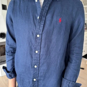 Mörkblå skjorta från Polo Ralph Lauren - 14-16 år, L. Säljer en klassisk mörkblå skjorta från Polo Ralph Lauren med vit knappstängning och röd broderad logga på bröstet. Skjortan har lång ärm och är tillverkad i ett mjukt bomullstyg. Perfekt för en stilren look!