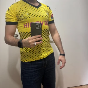 Borussia Dortmund gul fotbollströja - Gul kortärmad Borussia Dortmund fotbollströja med svarta detaljer och mönster av små svarta rutor. Svart V-ringad krage och svarta muddar vid ärmslut. Tryck med klubbmärke, sponsor och nummer 18 på ryggen tillsammans med texten Dortmund.