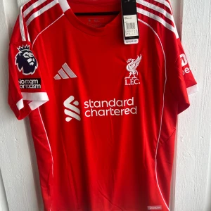 Liverpool FC Mohamed Salah matchtröja - Röd Liverpool FC fotbollströja med vita detaljer, tryck av M. Salah och nummer 11 på ryggen. Adidas-logga och Premier League-märke på ärmen. Standard Chartered sponsortryck på bröstet. Kortärmad modell i lätt funktionsmaterial.