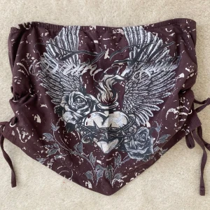 Brun bandeau topp med tryck från Urban Outfitters - Säljer en brun bandeau topp med coolt tryck av vingar, rosor och hjärta framtill. Från Urban Outfitters. Sidorna har snörning för justering 🧸Aldrig använd!