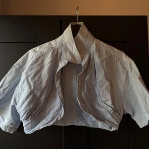 Ljusblå croppad bolero - Säljer en ljusblå croppad bolero med korta ärmar och klassisk krage. Skjortan har knappar framtill och en avslappnad passform, perfekt för en trendig och avslappnad stil.