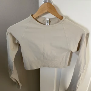 Beige croppad långärmad träningstopp från Lindex - Säljer en beige croppad långärmad träningstopp från Lindex i återvunnen polyamidblandning. Tröjan har rund halsringning och inbyggda uttagbara kupor. Perfekt för en trendig och sportig look.