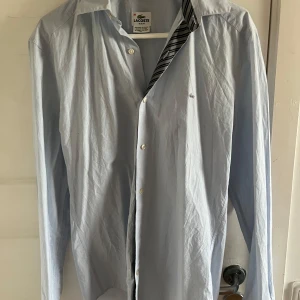 Ljusblå skjorta från Lacoste - Säljer en stilren ljusblå skjorta från Lacoste i slim fit-modell. Skjortan har klassisk krage, knappar framtill och diskret Lacoste-logga på bröstet. Perfekt för dig som gillar en enkel men snygg look. Använd 1 gång så nästan helt ny 