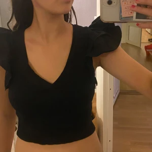 Svart croppad topp med volangärmar - Säljer en svart croppad topp med v-ringning och stora volangärmar. Toppen är tight och har en ribbad struktur som ger en snygg passform. Pris kan diskuteras 