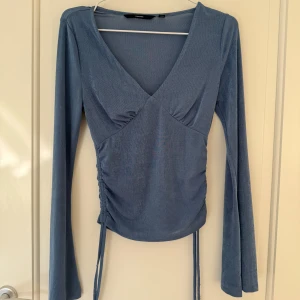 Blå långärmad topp med v-ringning från Vero Moda - Säljer en blå långärmad topp från Vero Moda med v-ringning och knytband i sidan. Toppen har en figurnära passform och är tillverkad i ett semi glittering material.