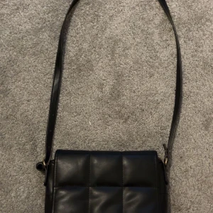 Svart quiltad axelväska - Snygg svart axelväska med quiltad design och justerbart axelband. Väskan har ett stort huvudfack och innerficka med dragkedja. Perfekt för att bära dina viktigaste saker med stil.