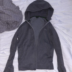 Grå hoodie med dragkedja från BOSS - Säljer en stilren grå hoodie från BOSS med dragkedja framtill och huva. Jackan har två fickor på sidorna, varav en med dragkedja, samt diskret BOSS-logga på bröstet. Perfekt för en avslappnad look.