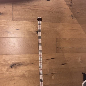 Rutig och svart bälte med metallspänne från Burberry - Ett bälte med två sidor från Burberry säljes. Ett i klassiskt beige rutigt mönster med svart metallspänne och ett i svart med diskret rutmönster och svart metallspänne. Båda har Burberrys logga på spännet och präglad häst på lädret.