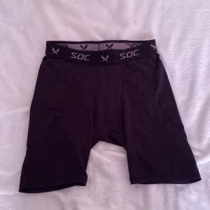 Svarta träningsshorts från SOC - Svarta, tighta träningsshorts från SOC med elastisk midja och logga runt linningen. Perfekta för gymmet eller löpning. Tillverkade i syntetmaterial som andas och ger bra rörelsefrihet.